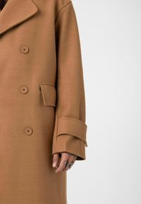 Wollen jas in kameelkleur met drie knopen, een klepzak en een riem aan de mouwmanchet; hand met ringen en tatoeages zichtbaar.