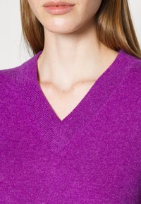 Pull à col en V de couleur violet vif, fabriqué en matière tricotée douce. Dispose d'un col côtelé et d'une texture subtile sur toute la surface.