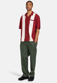 Camicia a maniche corte rossa e bianca con taschino frontale, abbinata a pantaloni larghi verdi. Design con bottoni e strisce verticali a contrasto.