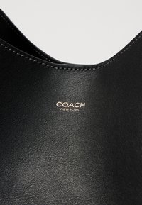 Sac en cuir noir avec une texture lisse, des coutures discrètes et un logo "COACH NEW YORK" embossé en or à l'avant.