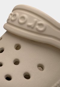 Κοντινό πλάνο μπεζ λαστιχένιας κλογκ με οπές αερισμού και μαλακό ιμάντα με ανάγλυφα μερικά γράμματα του ονόματος της μάρκας.