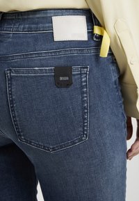Jeans de mezclilla azul oscuro con un acabado texturizado, que presentan un parche de cuero blanco y una etiqueta de tela negra en el bolsillo trasero. Cinta de acento amarilla visible.