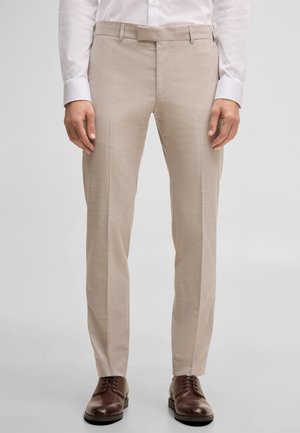 Man draagt beige op maat gemaakte broek, wit overhemd en bruine leren nette schoenen, staand tegen een effen lichte achtergrond.