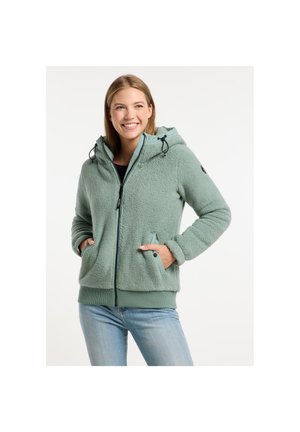 Femme souriante portant une veste polaire zippée vert clair avec capuche, les mains dans les poches, associée à un jean bleu, debout sur un fond blanc.