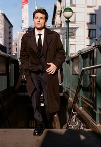 Mladý muž v tmavém trenčkotu a obleku stoupá po schodech metra v městském prostředí s budovami a dopravními značkami v pozadí.