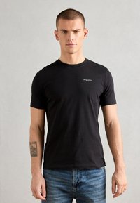 Tricou scurt din bumbac negru cu mânecă scurtă și guler rotund, având un mic logo alb pe partea stângă a pieptului. Textură netedă, design ajustat.
