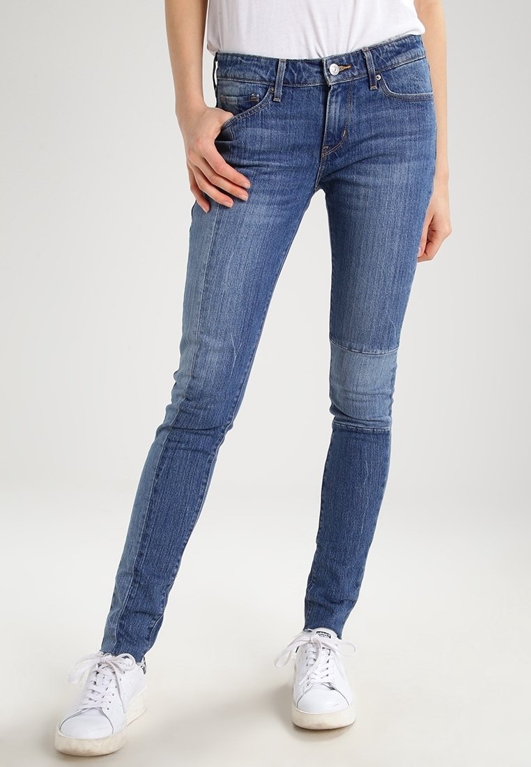 Personne portant un jean skinny patchwork bleu et des baskets blanches, debout avec une main dans la poche sur fond uni.