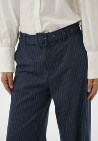 Fehér, hosszú ujjú blúz hímzett mandzsettákkal, amelyet sötétkék csíkos nadrággal párosítottak, hozzá illő övvel és fémlyukakkal.