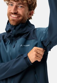 Veste Vaude bleu foncé avec un col montant, des poches zippées et des poignets ajustables. Texture lisse et coupe ergonomique pour le confort.