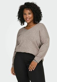 Femme aux cheveux bouclés portant un pull beige à col en V et un pantalon noir, souriante et debout devant un fond clair uni.