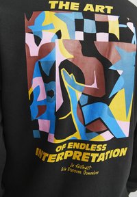 Sudadera negra con un diseño abstracto multicolor que presenta formas en azul, rosa, marrón y amarillo. El texto incluye "EL ARTE DE LA INTERPRETACIÓN INFINITA."