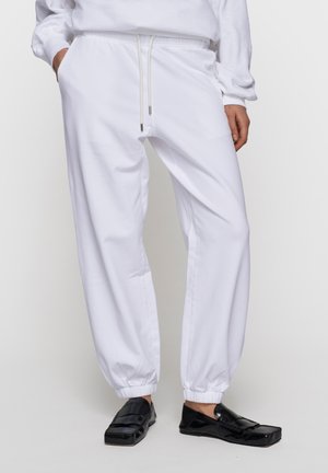 Persona che indossa pantaloni jogger bianchi con coulisse e mocassini neri lucidi, in piedi con le mani parzialmente nelle tasche.