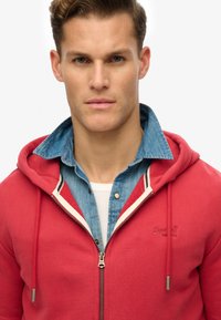 Felpa rossa con zip e cappuccio regolabile, indossata sopra una camicia di jeans azzurri. Materiale in cotone, presenta ricamo del logo e dettagli a contrasto.
