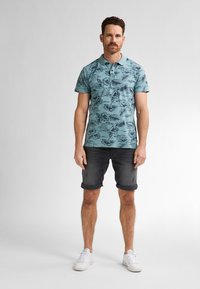 Světle modrá polo tričko s černým květinovým vzorem, krátké rukávy a límec. Kombinováno s srolovanými tmavými šortkami a bílými teniskami.