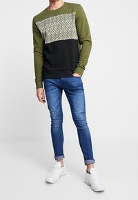 Sudadera verde con una sección superior estampada, sección inferior negra, combinada con jeans ajustados azules y zapatillas blancas.