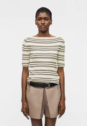 Femme portant un pull crème à manches courtes avec des rayures horizontales noires, un short plissé beige et une ceinture noire, debout devant un fond blanc.