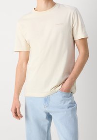 Camiseta de algodón beige con mangas cortas y escote redondo, que presenta un pequeño logo de Calvin Klein en marrón claro en el lado izquierdo del pecho.
