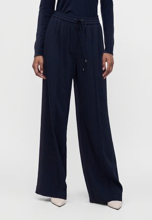 La personne porte un pantalon large taille haute bleu marine avec une taille à cordon et des chaussures blanches pointues, debout devant un fond uni.