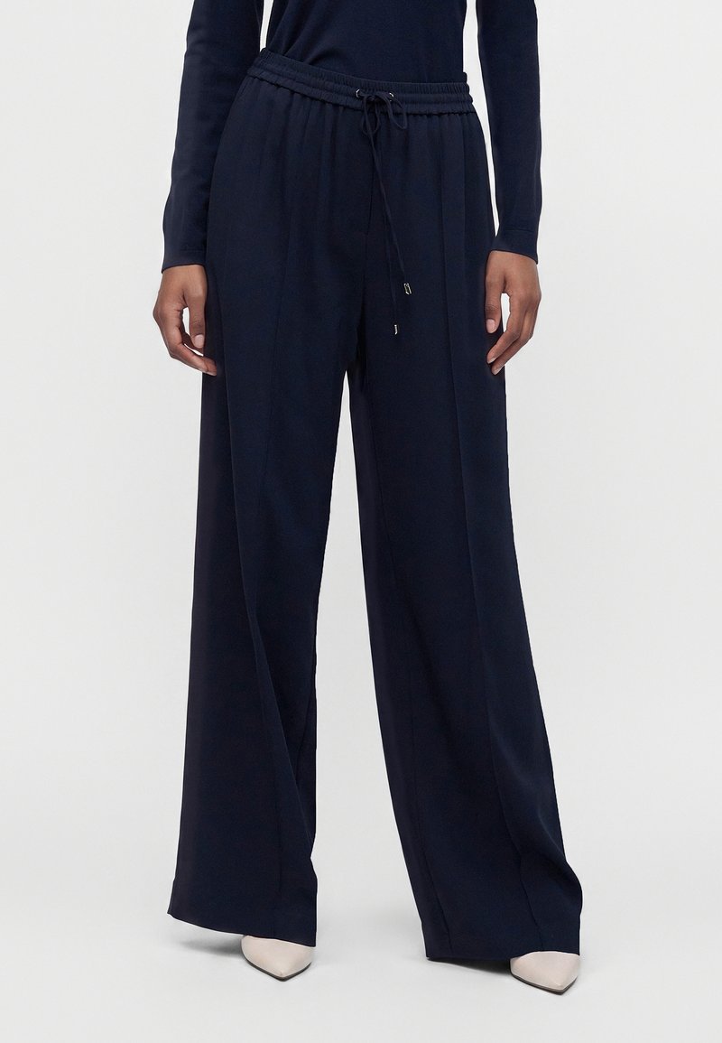 La personne porte un pantalon large taille haute bleu marine avec une taille à cordon et des chaussures blanches pointues, debout devant un fond uni.