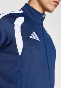 Αθλητικό μπουφάν ναυτικού μπλε με λευκές λεπτομέρειες, ψηλό γιακά και κούμπωμα με φερμουάρ. Κατασκευασμένο από υφή υφάσματος με πάνελ από δίχτυ στα μανίκια.