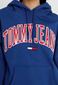 Sudadera con capucha azul con el texto en blanco y rojo "TOMMY JEANS" en forma de arco. Incluye una pequeña franja de logo en rojo, blanco y azul en el bolsillo.