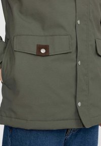 Olivgrön jacka i slätt tyg, med en knappad ficka med bruna mockadetaljer, tryckknappar och en rak nederkant.