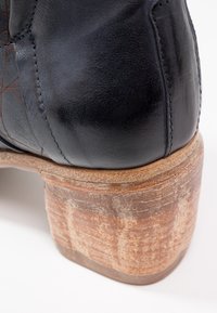 Gros plan du talon d'une chaussure en cuir noir avec couture visible et semelle en bois superposé brun texturé sur fond blanc.