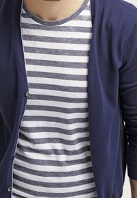 Marinblå cardigan i mjuk stickad kvalitet, med knäppning. Bärs över en figurnära randig t-shirt i grått och vitt. Slät textur.