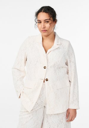 Frau mit lockigem Haar trägt ein cremefarbenes Spitzenblazer und passende Hose, steht mit einer Hand in der Tasche vor weißem Hintergrund.