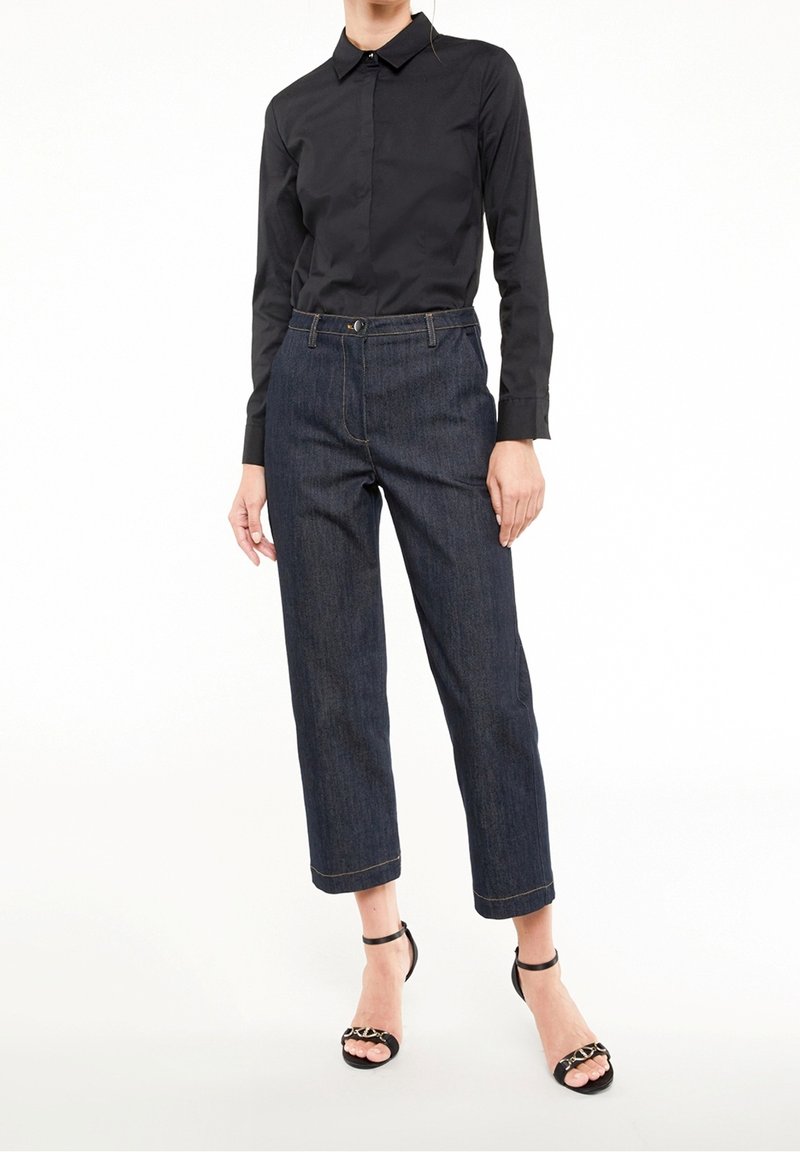 Chemise noire à manches longues, pantalon en denim foncé raccourci avec couture visible, et sandales noires à brides. Lignes épurées et coupe ajustée.
