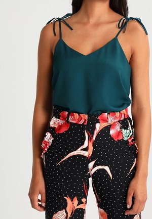 Camisola de seda color azul verdoso con tirantes finos que se anudan en los hombros, combinada con pantalones negros de cintura alta con un patrón de lunares y detalles florales rojos.