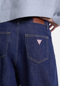 Jeans in denim blu scuro con cuciture gialle a contrasto, dotati di una patch in pelle in vita e di un logo rosso triangolare sulla tasca posteriore.