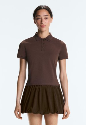 Nő barna rövid ujjú galléros pólót és hozzá illő barna fodros miniszoknyát visel, egyszerű világos háttér előtt áll.