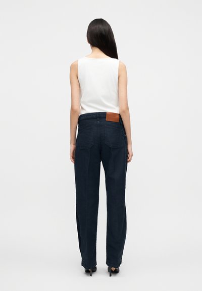 Ženska z dolgimi temnimi lasmi, oblečena v belo majico brez rokavov, mornarsko modre široke hlače in črne čevlje z visokimi petami, stoji pred enobarvnim ozadjem.