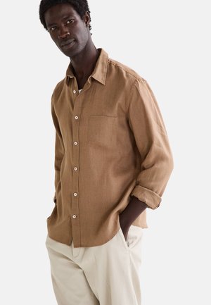Chemise boutonnée marron à manches longues en tissu texturé, dotée d'une poche poitrine unique et de boutons blancs, associée à un pantalon de couleur claire.