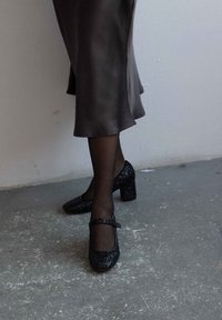 Černé třpytivé Mary Jane podpatky s chunky podpatkem, kulatou špičkou a páskem přes nárt, doplněné o průhledné černé punčochy a šaty bez ramínek.