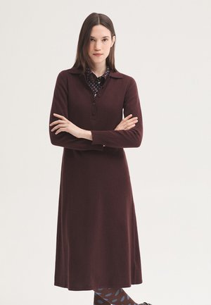 Jeune femme aux cheveux bruns et raides portant une robe bordeaux à manches longues et des collants à motifs, se tient les bras croisés sur fond blanc.