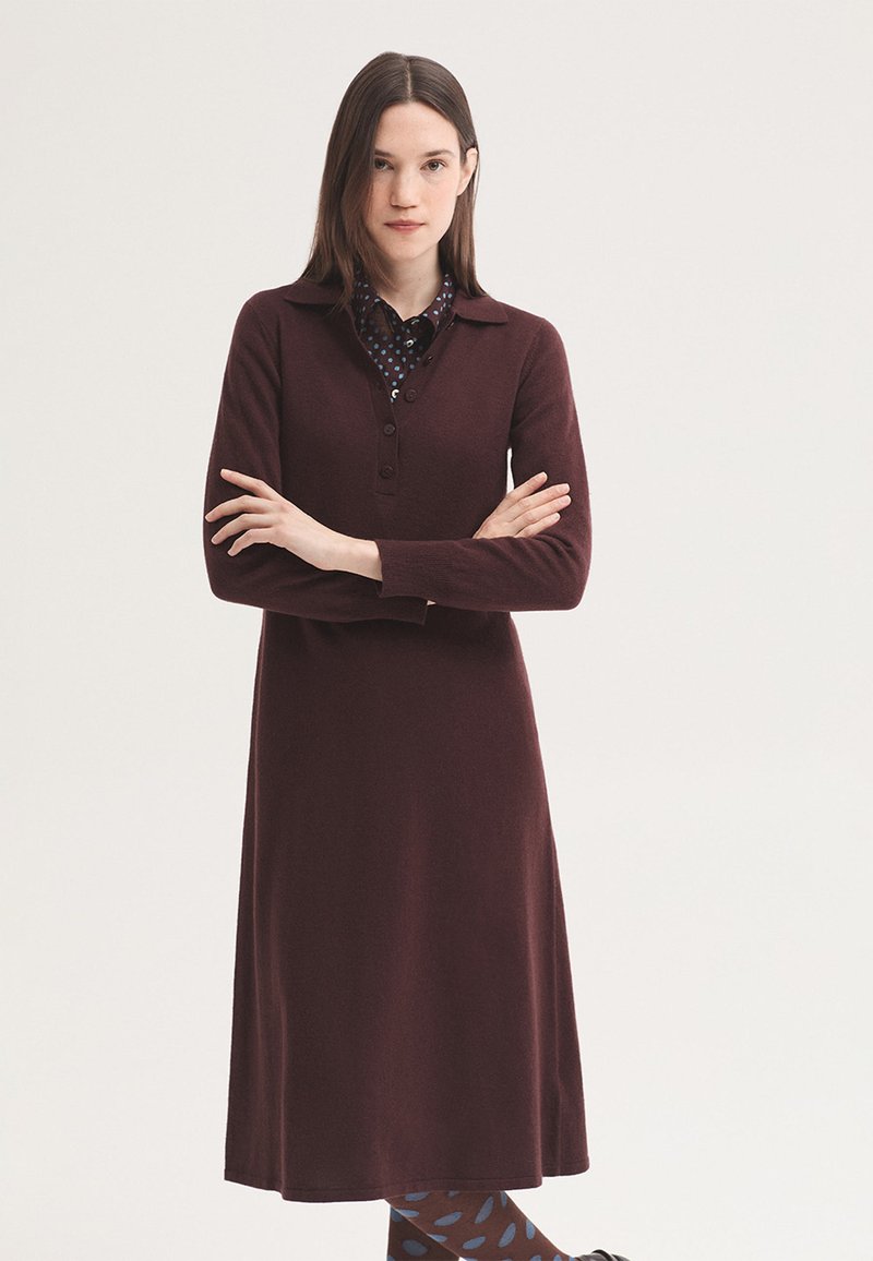 Jeune femme aux cheveux bruns et raides portant une robe bordeaux à manches longues et des collants à motifs, se tient les bras croisés sur fond blanc.