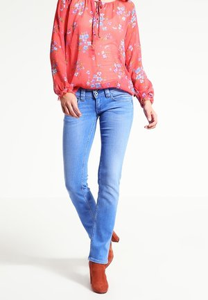 Jean en denim bleu clair à coupe slim, associés à un blouse rouge à motifs floraux avec manches longues et décolleté à cordon. Bottines brunes.
