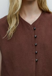 Brun ärmlös plagg med v-ringning, med fem svarta knappar fram. Tyget verkar texturerat med ett subtilt vävmönster.