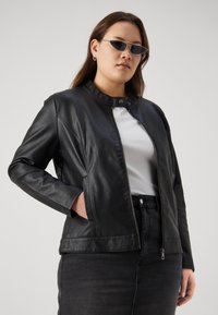 Black leather jacket with a stand collar, front zip closure, and side pockets; worn over a white shirt and black denim skirt.