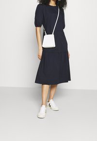 Mörkblå bomullsklänning med figursydd överdel, puffärmar och lager-på-lager kjol. Kombinerad med en vit axelremsväska och vita sneakers.