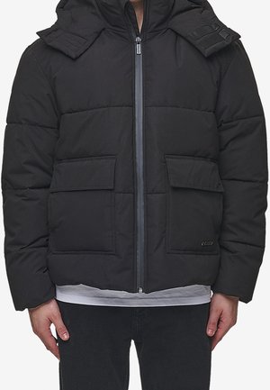 Sort polstret jakke med lynlås foran, to lommer med klap og fastmonteret hætte, båret over en hvid skjorte og sorte bukser.