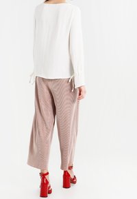 A woman wears a white long-sleeve top and wide pink trousers, showcasing red heeled sandals. She is viewed from the back against a white backdrop.