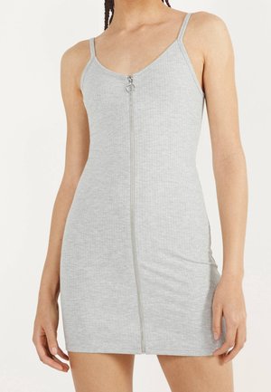 Mujer vistiendo un vestido ceñido sin mangas de color gris claro con tirantes delgados y una cremallera plateada en la parte frontal que va desde el escote hasta el dobladillo.