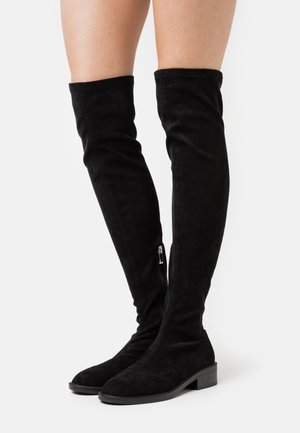 Bottes noires en suede au-dessus du genou avec une fermeture éclair sur le côté, semelle plate et texture lisse, conçues pour un ajustement près du corps.