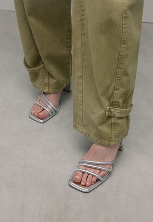 Silberne Sandalen mit mehreren dünnen Riemen und quadratischer Zehenpartie, kombiniert mit olivgrünen Hosen, vor einem strukturierten grauen Boden.