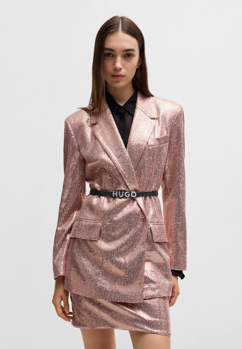 Blazer lucido color oro rosa con gonna coordinata, caratterizzato da un design strutturato, tasche e una cintura nera con logo "HUGO". Camicia nera con colletto sotto.