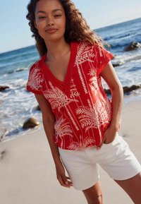 Top corto rojo de manga corta con patrones de helechos blancos y escote en V, combinado con pantalones cortos blancos, de pie en una playa cerca de las olas del océano.