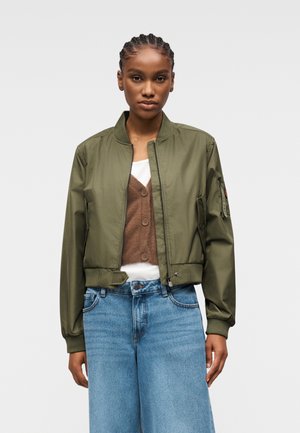 Giovane con capelli intrecciati che indossa una giacca bomber verde oliva aperta, gilet marrone con bottoni, camicia bianca e jeans azzurri chiaro su sfondo semplice.